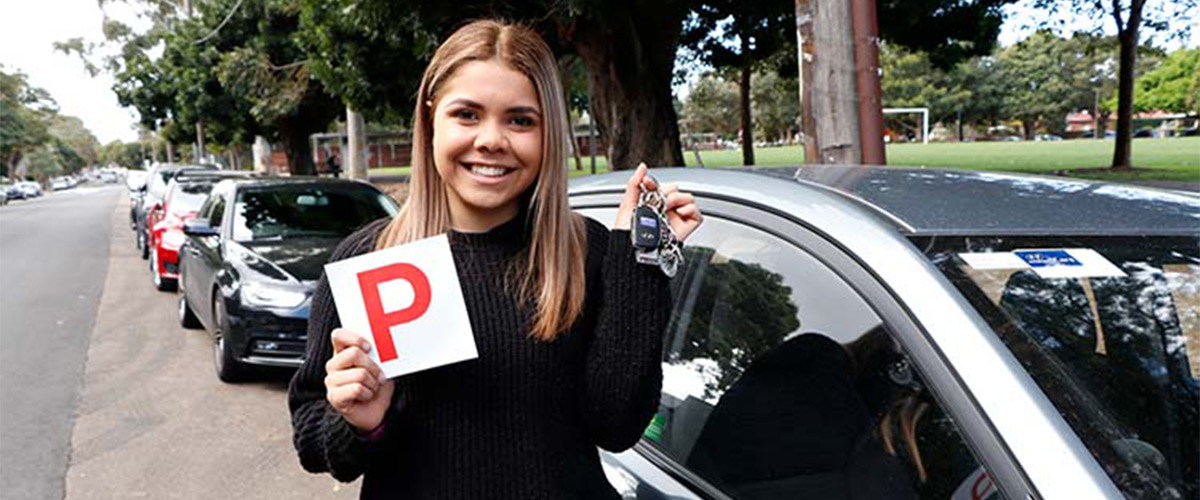 Woman holding up a white and red P-plate sign, standing next to a car.